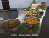A buffet spread of Fusion Asian dishes in stainless steel chafing dishes on a white tablecloth, with greenery decorations and a beverage dispenser in the background.